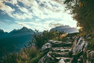 Rocky mountain trail under a partly cloudy sky with shrubbery on either side and peaks in the distance.