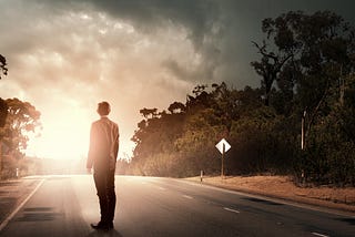 A man dressed in a suit stands on a road looking to the sunrise in the distance.