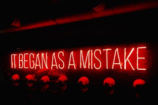 We see a neon red sign saying “It began as a mistake”.