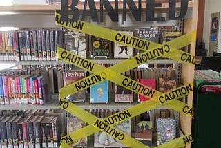 Image of library bookshelf covered in caution tape with the word BANNED at the top in black construction paper letters.