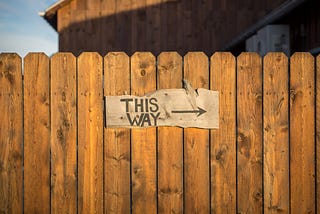 A rustic fence made of brown wood holds a sign that says “THIS WAY” with an arrow pointing to the right.