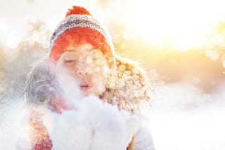 Young white woman in a snowy field wearing winter pompom hat, warm hooded ski jacket, blowing snow from white fluffy mittens