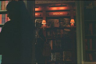 picture of a woman on the left side, looking to the left at some books. In the background, another woman is browsing bookshelves in a dimly lit room.