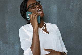 A woman in a white blouse is holding her phone laughing. The other hand is clasping her stomach.