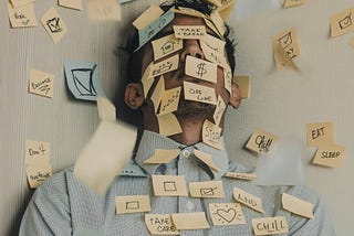 A man in business attire stands against a wall with his face, shoulders and the wall covered in reminders on sticky notes.