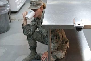 J L Cross enjoying a cup of coffee while she was deployed in Kuwait in year 2017. She’s seen dressed in her AF Airman Battle Uniform sipping from a large white mug. She’s hunkered under a steal kitchen table, an attempt to avoid leadership at the time. Shown in the back ground is the Kitchen in which she worked to serve hundred, if not during some rotations of manning, over a thousand Marines, Navy, Air Force, Army, and other allied international forces at her base.