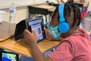 A masked school-aged child wearing headphones looks at a tablet device while sitting at a school desk.