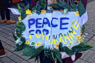 Colour photo. An unknown person holds a hand-made cardboard banner which reads ‘peace for Ukraine’.