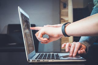 An open laptop, photographed from the side. A hand is pointing at the screen. Another hand is navigating the laptop’s mouse.