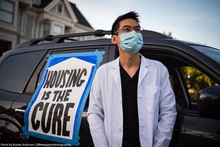 A person in a medical mask and white doctor’s coat stands in front of a vehicle with a sign that reads “HOUSING IS THE CURE”