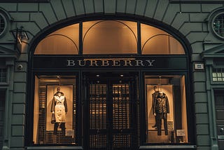 The storefront of a Burberry shop on the high street, closed for the evening, with fashion offerings softly lit from behind in the window.
