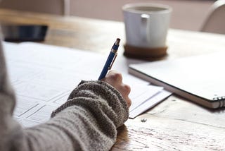 Stock photo of a person in a gray sweater with a pen or mechanical pencil writing on documents while sitting at a desk. A notebook and a cup of coffee are also on the desk.