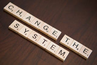 Scrabble tiles spelling out “change the system” sit on a wooden table.