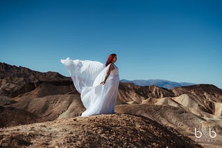 A woman dressed in a white flowing dress, which is blowing in the wind. She is standing on a rocky hill in desert terrain.