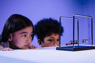 two children stating at a Newton’s cradle or pendulum — a device that typical sits on desktops, consisting of five metal hanging balls. when one ball is pulled sideways then released, it collides with the other balls, which forces the ball on the other side into motion, thus starting a back-and-forth chain reaction that, while not literally “perpetual,” tends to continue for a very long time.