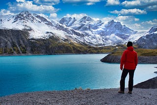 A blue lake nestled beneath a snow-capped mountain with husband standing on the side of the lake watching and mesmerizing the beautiful day in nature