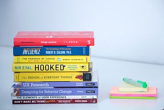 Vários livros de design empilhados, ao lado dos livros alguns post-its coloridos e uma caneta marcadora na cor verde.