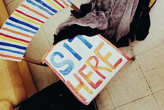 Bird’s eye view of wooden and metal stack chair painted in white, red, orange and blue, with text on seat: “SIT HERE”.