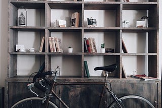 Photo of a gray bookshelf with books sparsely spaced and a road bicycle leaning in front of it. Neutral toned and relaxing to look at.