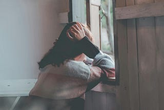 A woman leans against a windowsill in quiet contemplation, resting her head on her hand. Soft light filters through an open wooden window, creating a calm, introspective atmosphere.