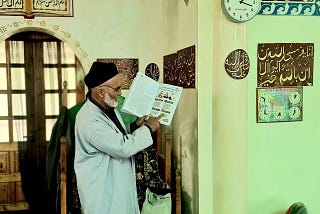 Domullo Shermuhamad stands at the front of the mosque reading from a Healthy Mother, Healthy Baby booklet.