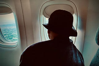 An elderly woman wearing a hat and looking out a window of a plane descending for landing.