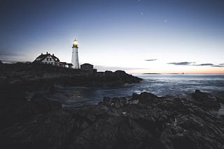 A lighthouse exudes a soft beam as dusk approaches against the backdrop of a dark, rocky ocean.