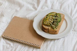 Aesthetic stock image shows avocado toast on a plate on a bed with white sheets next to a brown hardback notebook. The image is purely for decorative use only to support the blog post on Defining Wellness; The 6 Core Pillars of Health and Wellness and How To Practice Them