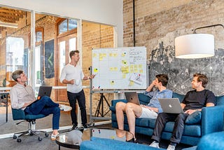 A group of people is gathered in a room, actively engaged with their laptops and collaborating around a whiteboard.