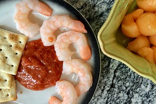 shrimp cocktail with saltines and cocktail sauce on a plate on a gray marble counter next to a green bowl of canteloupe balls