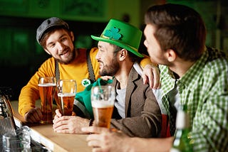 A group of three young men sit at a bar with pints of beer at the ready. They are smiling and obviously having a good time. Two are wearing hats; one of the hats is bright Irish green and sports a shamrock.
