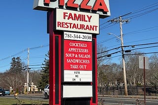 The photo shows a free-standing sign in front of Town Spa, a restaurant in Stoughton, Massachusetts. Town Spa is at the top in white letters, below it is pizza spelled out in red. The bottom part of the tall sign lists other food items and their telephone number.