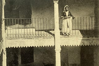 1912 black and white postcard of a two storey moorish house with proches. A woman stands on the second floor balcony. She is wearing a traditional costume with a short jacket and flowing skirt