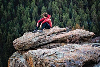 young man wearing red jacket and black pants sitting on boulder looking out toward forest