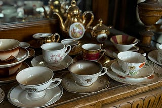 Photograph of multiple white and painted china teacups sitting on china saucers on a brown wooden sideboard, with a gold teapot in the background.