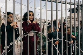 Palestinian women & children peer through the bars of yet another randomly constructed Israeli choke point in Hebron