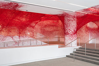 Standard white walls and concrete floor of a modern museum lobby. Stairs are to the right and there’s a central column. Along the walls, ceiling, and even the column is red yarn, like a massive, messy spiderweb.