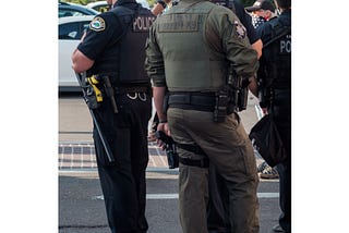 Two police officers, center frame, stand facing away from the camera. One has his hand on gun.