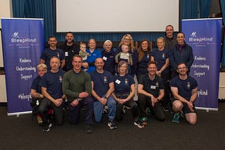 16 people in a group shot (two rows, front row are kneeling) made up of MDP fitness instructors, Ali, and Carl and Jen with their babies, either side is a BleepKind roll up banner and there is a projector screen in the background