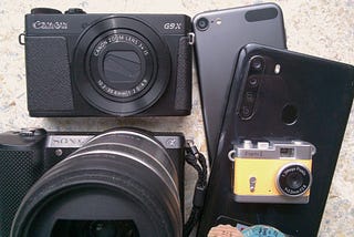 group of digital picture-taking devices: 2 black smartphones and orange ‘toy camera’ (right), as well as dSLR and enthusiast pocket camera (left)