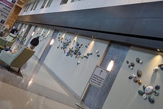 Hospital entrance hall with blue-gray walls adorned with smooth natural-colored clustered stones and recessed lighting and a sign pointing ahead for admissions or radiology.