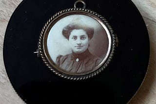 An antique brooch from the early 1900’s with a photo of a Scots-Irish woman with dark hair and dark eyes looking at the camera. She is wearing a black high-collared dress with puffed sleeves with a white brooch on her collar. Her hair is in a loose bun on top of her head. The brooch is on a black obsidian scrying mirror.
