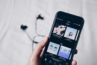 A hand with a smartphone inside where the Spotify app is opened. In the background earphones are lying on a sheet.