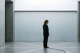 A single figure — gender neutral, mid-40s, sharp professional dress — standing in a vast empty space, architectural in feel. Concrete or glass. Six faint geometric lines or frequencies radiating outward from the figure — subtle, structural, not decorative. One of the lines is visibly interrupted — fractured mid-transmission. The figure is not in distress. They are still. The fracture is in the architecture around them, not in their expression. Lighting: cool, precise, slightly clinical. No text