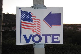 Photo displays a sign duct-taped to a pole with an American flag on one side and a left-facing arrow above the word “vote.”