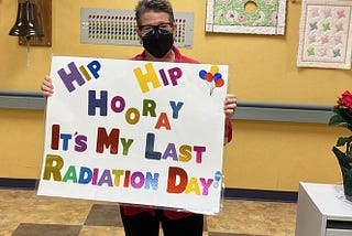 CC holding up a sign that reads, Hip Hip Hooray, It’s my last radiation day! Standing in front of the bell she is about to ring to celebrate the end of radiation!