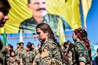 A group of revolutionary women in Rojava are gathered under a flag that shows Abdullah Öcalan