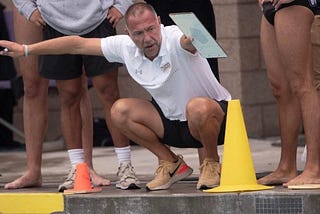 Photo of Coach Preslav Djippov crowched down in front of a yellow cone, and the edge of the pool. He is wearing a white polo shirt, black shorts, and running shoes. He is surronded by the legs of swimmers.