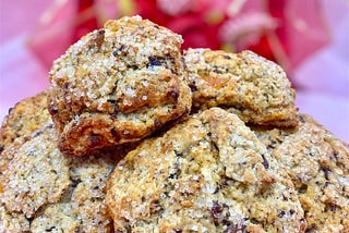 Scones flecked with chocolate and sanding sugar, stacked with a soft white background with blurred red and gold ribbon and pink and red flowers. Inspired by Lady Danbury