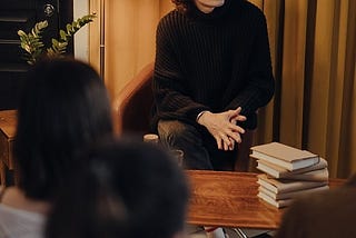 A young man (author) sitting in front of a pile of books on the table. He seems to have a little meeting or a chat with a few individuals who maybe are giving feedback about the book.
